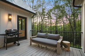 Una terraza de madera con una parrilla, un sofá y vistas a un bosque.