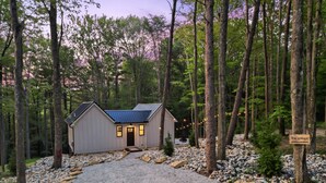 A cabin with a gravel driveway, surrounded by trees and a sign reading "The U Haus". - The U Haus | Hocking Hills, Modern & New, Hot Tub (Rockbridge)