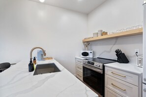 A compact kitchen with a sink, microwave, oven, and a shelf with dishes.