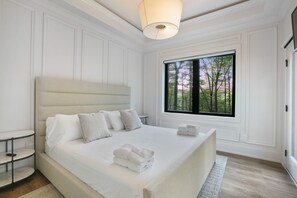 Una cama grande con sábanas blancas y varias almohadas, una mesita de noche, una ventana con vistas a árboles, y un aplique de techo.