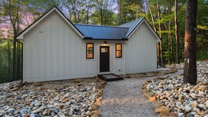 Une petite cabane blanche, avec un toit sombre, entourée d’un chemin de gravier et d’arbres.