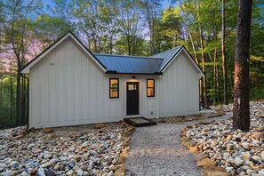 A small, white cabin with a dark roof, surrounded by a gravel path and trees.