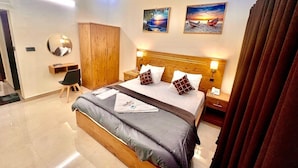 Una habitación de hotel moderna con una cama grande, cabecera de madera, mesitas de noche con lámparas, un armario, un espejo redondo y dos cuadros enmarcados en la pared.