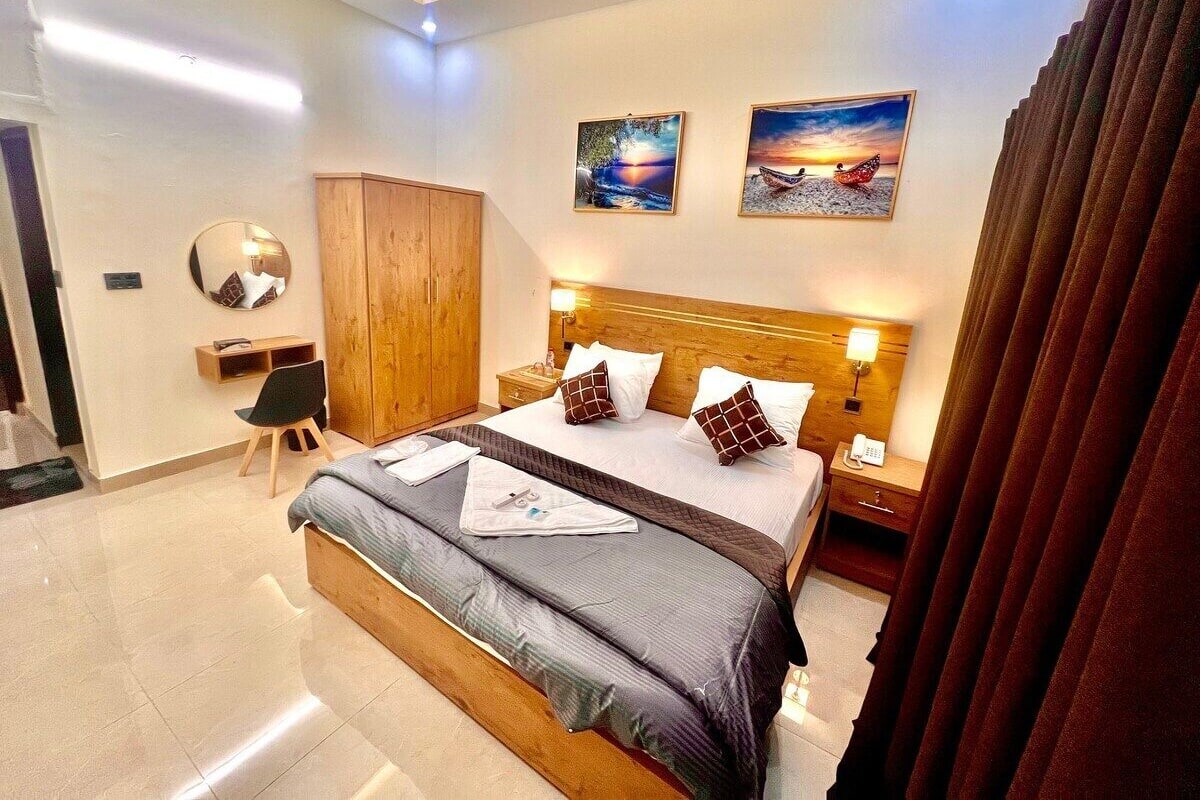 A modern hotel room with a large bed, wooden headboard, bedside tables with lamps, a wardrobe, a round mirror, and two framed pictures on the wall.