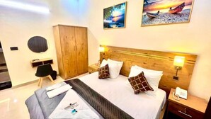 Una habitación de hotel con una cama grande, cabecera de madera, dos cuadros en la pared, un armario y una mesita de noche con lámpara.