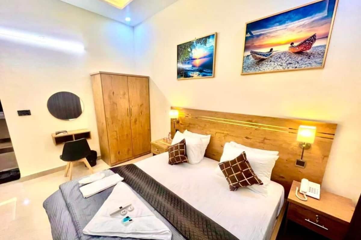 A hotel room with a large bed, wooden headboard, two paintings on the wall, a wardrobe, and a bedside table with a lamp.