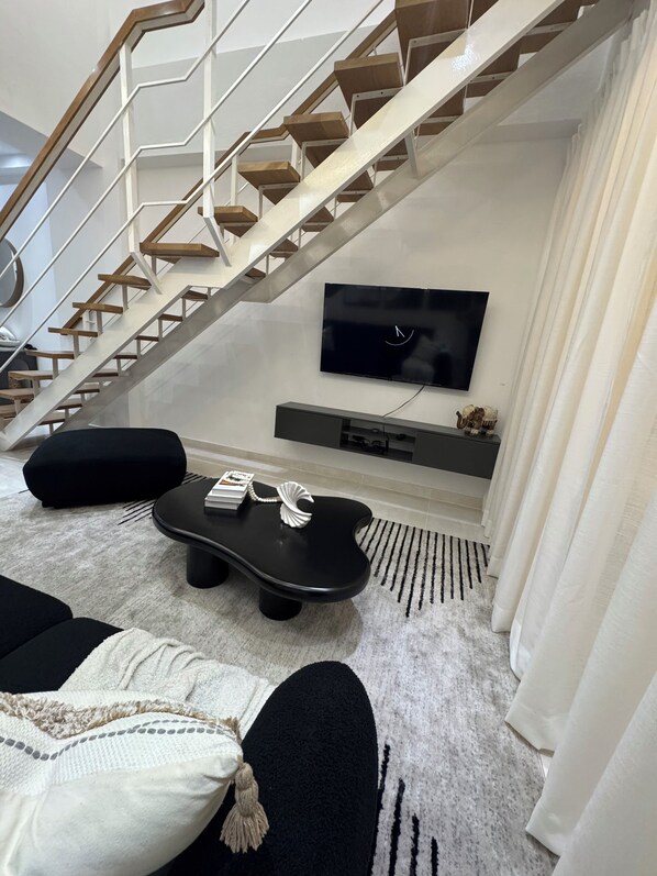 Una sala de estar moderna con una escalera, un televisor de pantalla plana, una mesa de centro negra y un sofá negro.