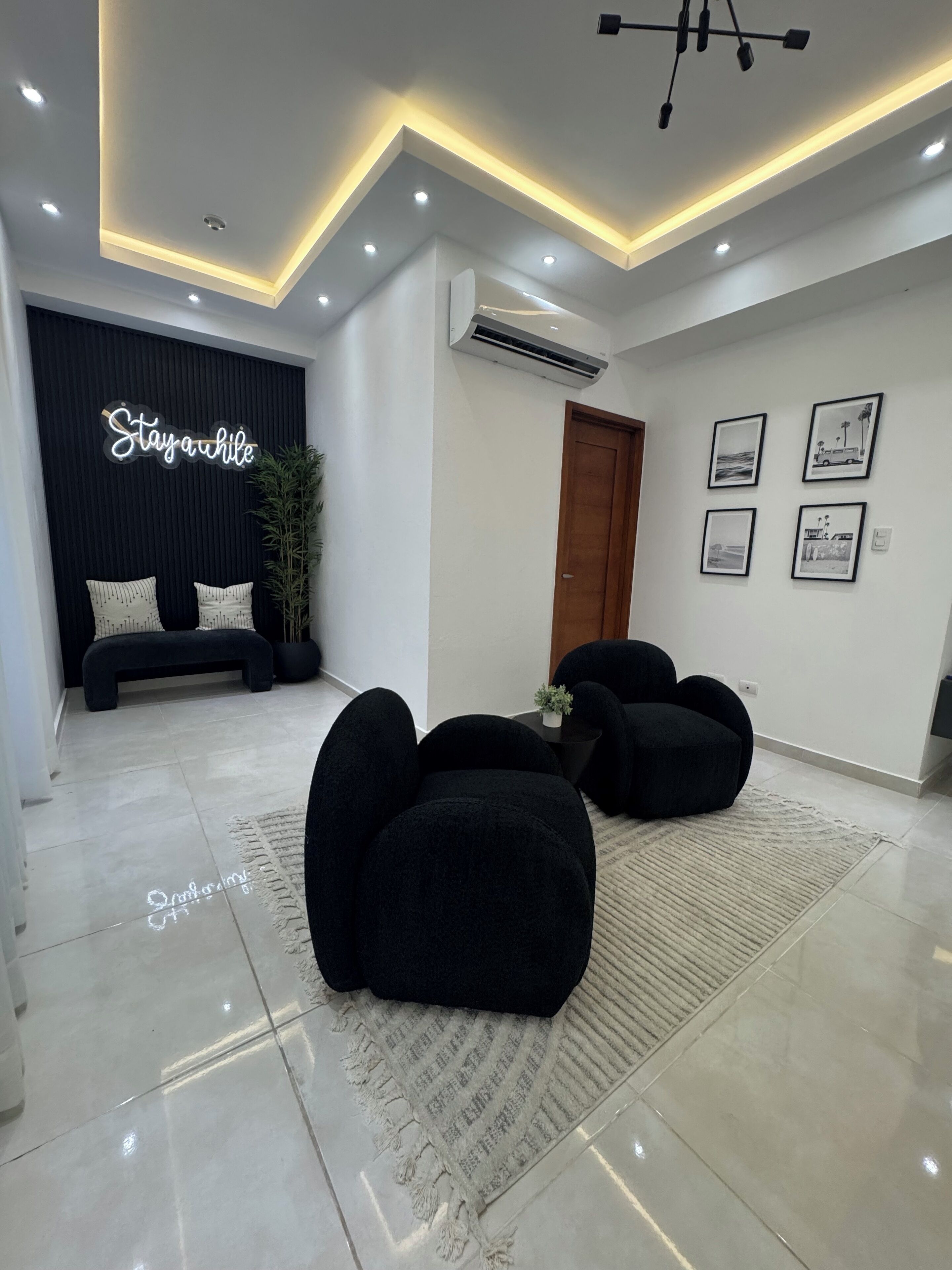A modern living room with a black sofa set, a black bench, and a rug with text. There is a wall-mounted air conditioner and a door.
