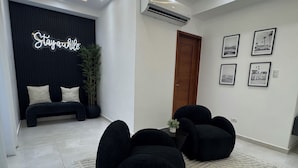 A modern living room with a black sofa set, a black bench, and a rug with text. There is a wall-mounted air conditioner and a door. - 3 bedrooms, 4 bathrooms, private pool and much more. (Playa Juan Dolio)