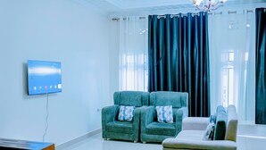 A modern living room with a flat-screen TV mounted on the wall, two green armchairs with patterned cushions, a beige sofa, and a large window with dark curtains. - Peaceful apartment with WiFi in charming Musanze-down town. (Musanze)