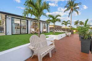 A modern outdoor patio with a wooden deck, white lounge chairs, and a well-maintained lawn. - Waterfront Duplex Tropical Oasis (Dania Beach)