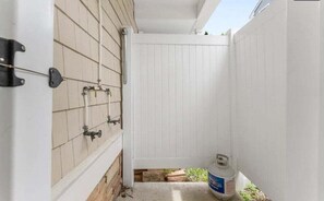 Un mur extérieur blanc, une porte en métal, une bouteille de gaz et un petit espace aménagé en jardin.