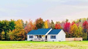 Une maison de plain-pied avec un porche, entourée d’un champ verdoyant et d’une forêt aux arbres aux couleurs automnales.