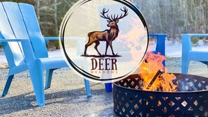 Un logo représentant un cerf et le texte « Deer Meadows » sur un panneau circulaire, placé entre deux chaises Adirondack bleues, avec un feu de camp dans un foyer métallique.