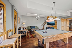 A pool table in a room with wooden flooring, a sofa, and a television.