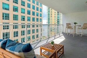 A balcony with a view of high-rise buildings, a wooden bench, a small table with a plant, and two chairs.