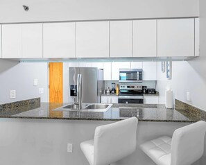 A modern kitchen with white cabinets, stainless steel appliances, granite countertops, and two white chairs. - Grand 1BR | Free Parking | Near Bayfront Park (Miami)
