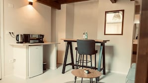 Una cucina compatta con macchina da caffè, forno a microonde e tostapane su un piano di lavoro bianco. Un piccolo tavolo con una sedia si trova al centro, e un quadro incorniciato appeso al muro.