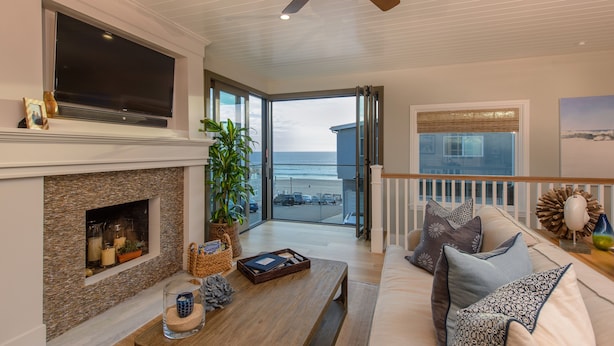 A spacious living room with a fireplace, a large flat-screen TV, a ceiling fan, and a balcony with a view of the beach.