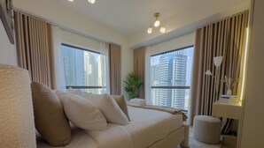 Una habitación de hotel moderna con una cama grande, dos ventanas con cortinas, una lámpara de noche, una mesita con un florero y vistas a edificios altos.