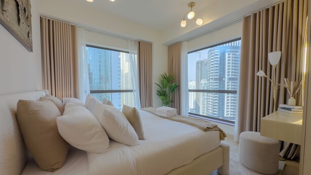 Una habitación de hotel moderna con una cama grande, dos ventanas con cortinas, una mesita de noche con lámpara y vistas a edificios altos.