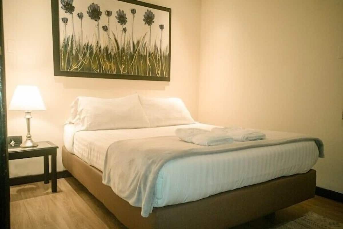 Una cama bien hecha con sábanas blancas, una mesita de noche con una lámpara, y un cuadro floral en la pared.