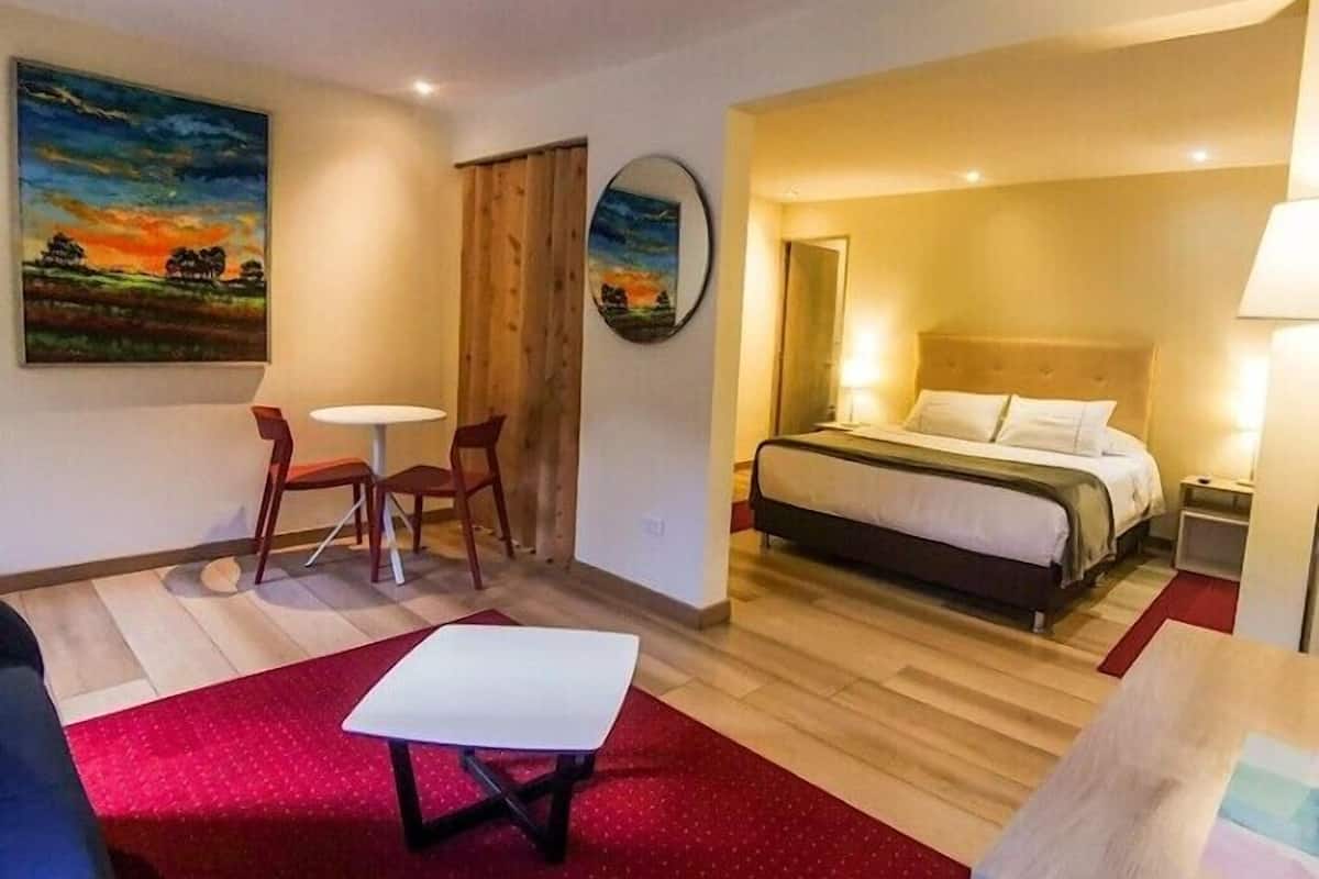 Una habitación de hotel con cama, una mesita, una silla, un escritorio y dos cuadros en la pared.