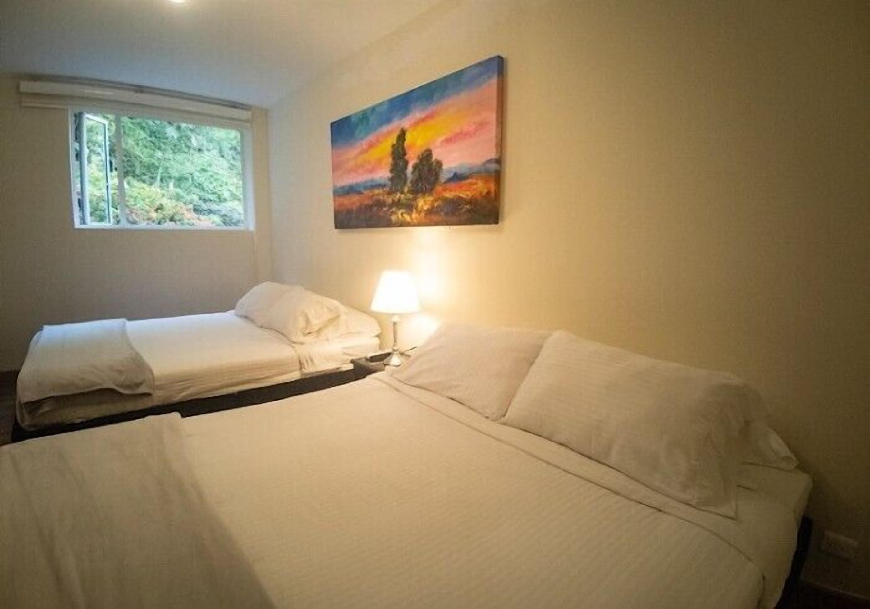 A hotel room with two beds, a painting on the wall, and a window with a view of greenery.