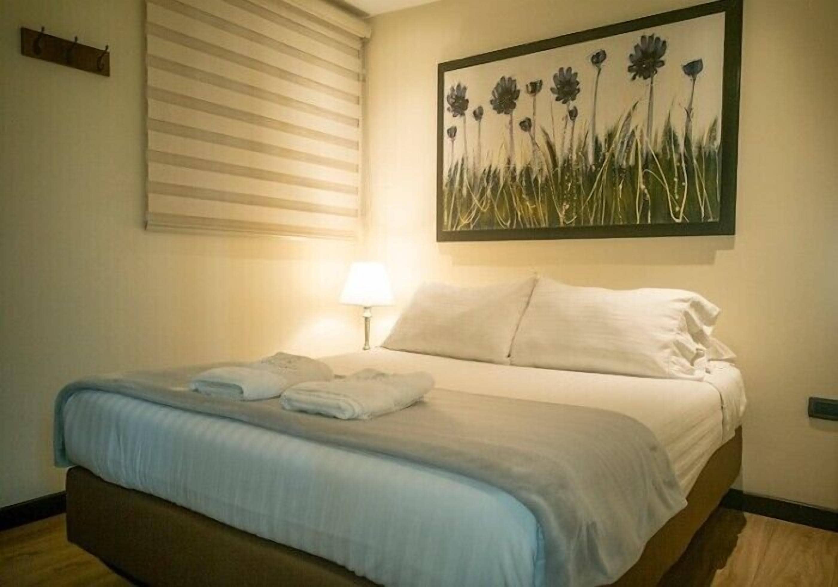 A neatly made bed with a blue blanket and pillows, a bedside lamp, and a painting of flowers on the wall.