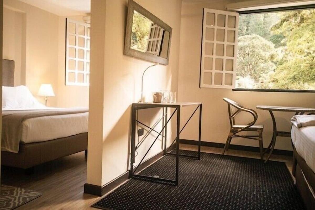 Habitación de hotel con cama, escritorio, silla, una mesita, espejo y ventana con vista a árboles.