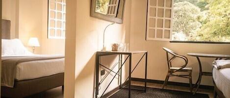 A hotel room with a bed, a desk, a chair, a small table, a mirror, and a window with a view of trees.