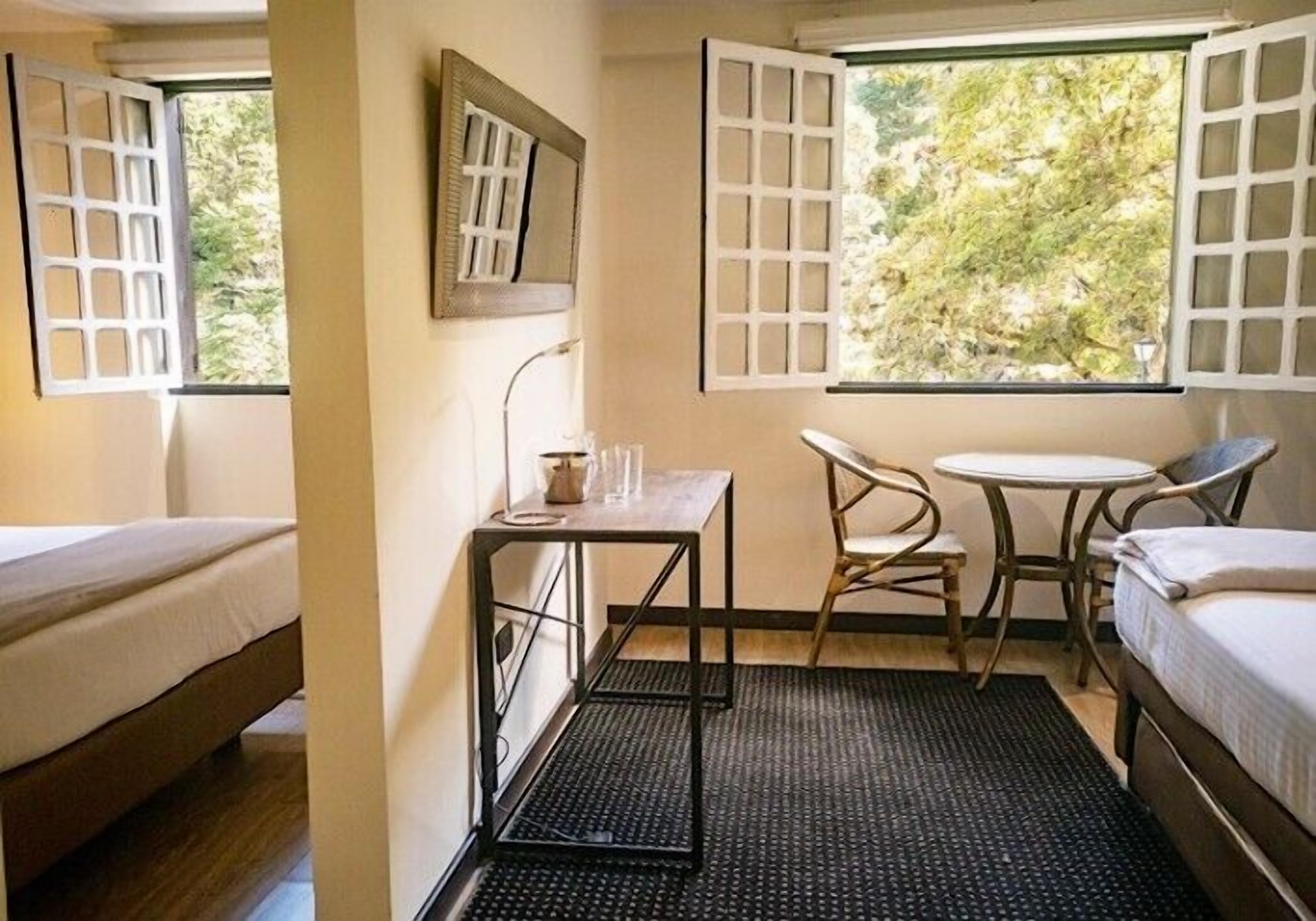 A room with two beds, a small table with a chair, and a window with open shutters.