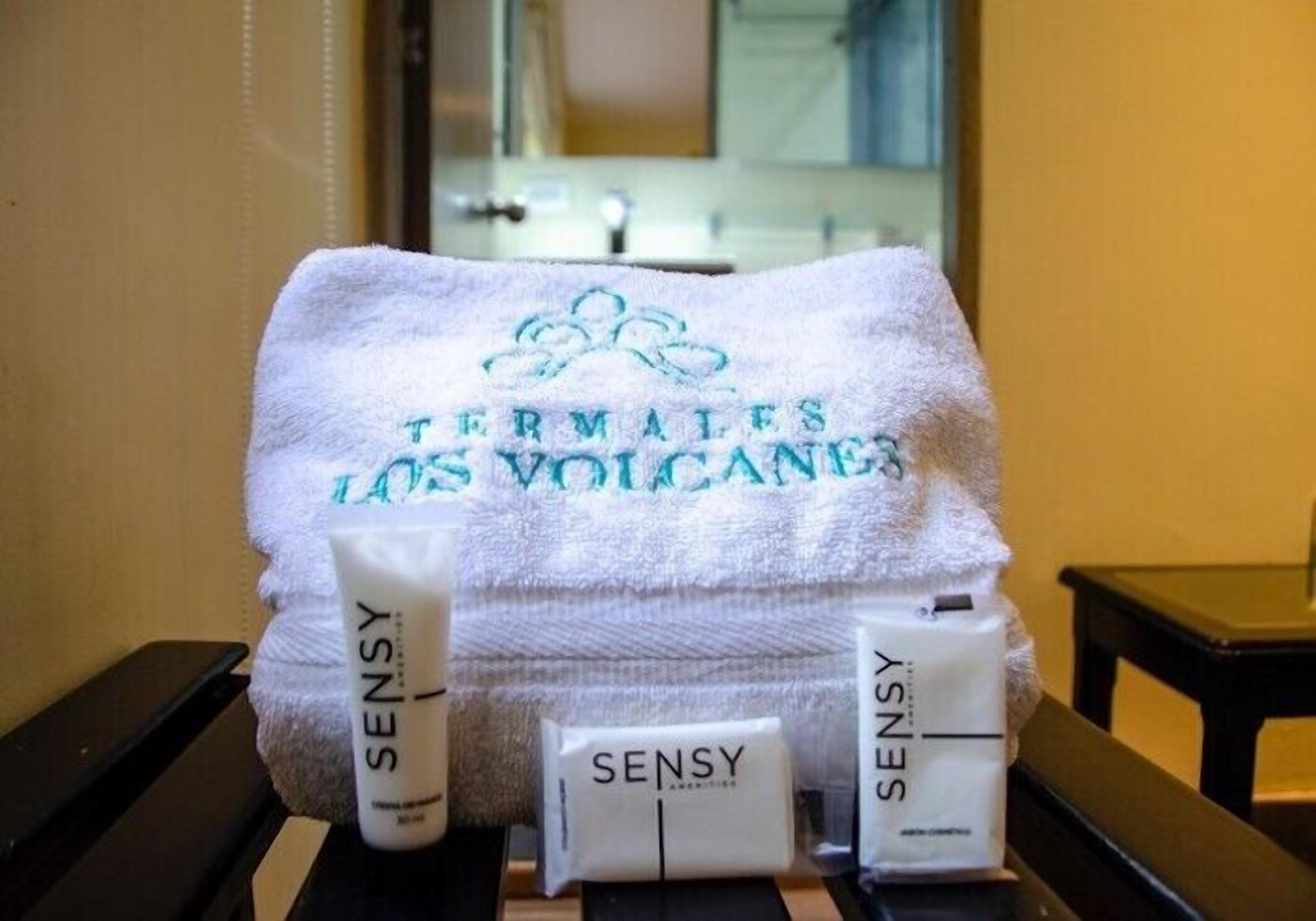 Un baño con un perchero que sostiene una toalla blanca con el bordado "Termales Los Volcanes", y tres botellas de jabón corporal Sensy.
