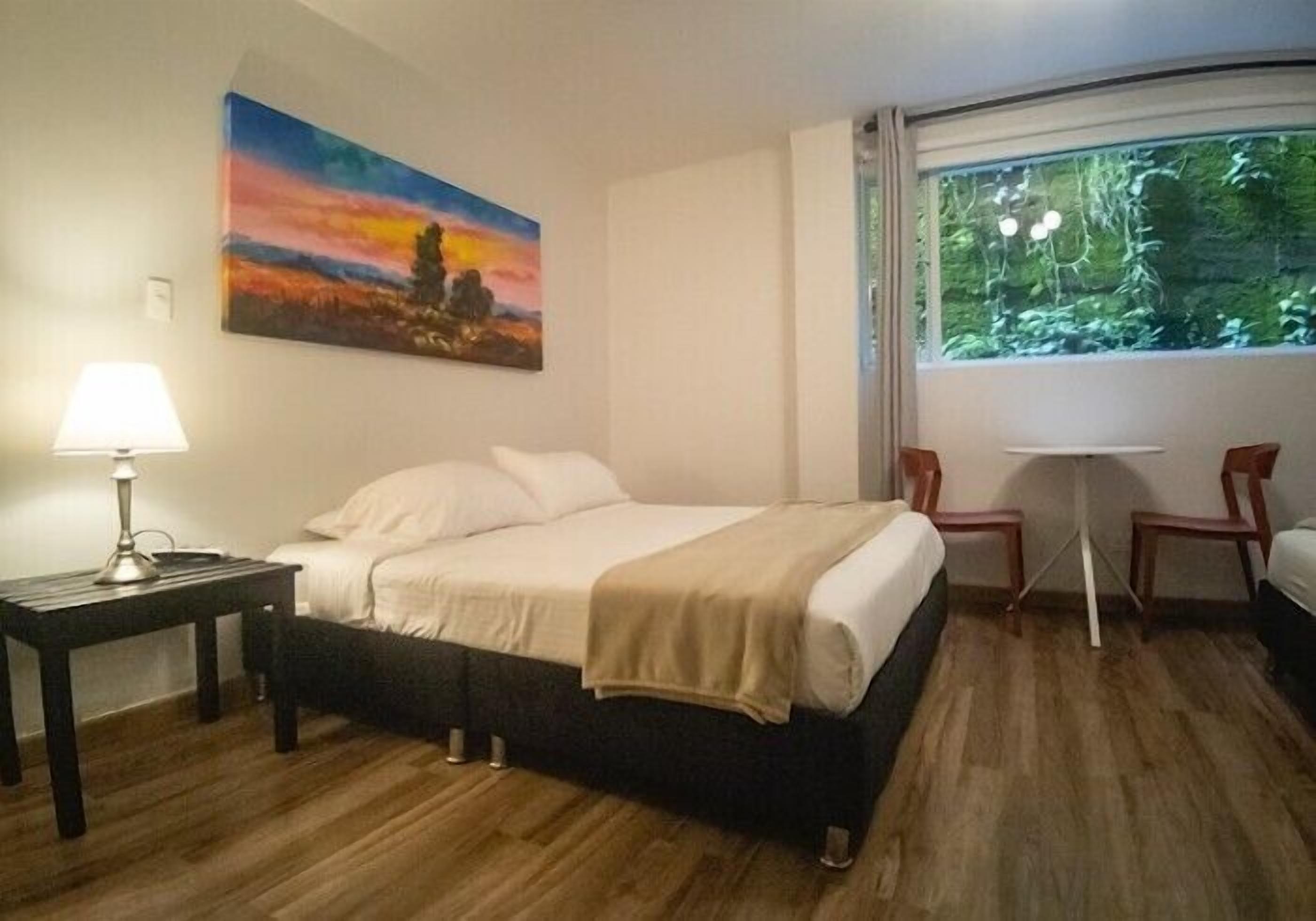 A hotel room with a bed, a desk with a lamp, a painting, and a view of greenery through a window.