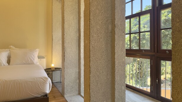 Ein Schlafzimmer mit Holzboden, einem Bett mit weißer Bettwäsche, einem Nachttisch und einem großen Fenster mit Blick auf Bäume.