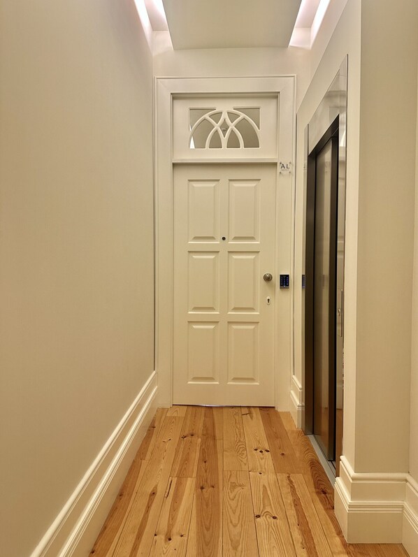 Un couloir doté d’un plancher en bois, d’une porte blanche ornée d’un motif décoratif en partie supérieure et d’un ascenseur fixé au mur.