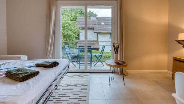 A bedroom with a bed, a small round table, a chair, and a view of the outside through a sliding glass door.