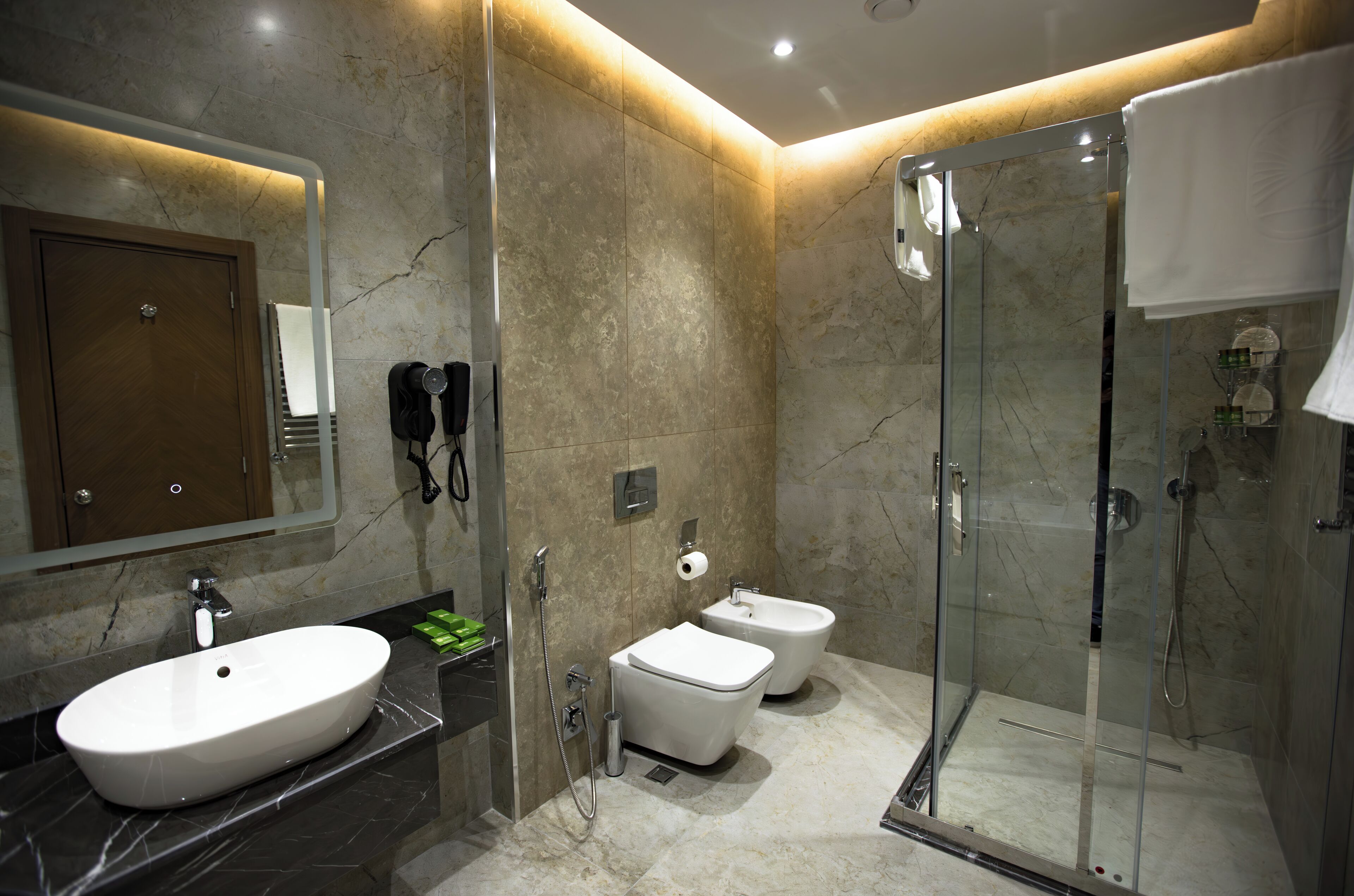 Un baño moderno con lavamanos de mármol, dos inodoros, una ducha con mampara de vidrio y un espejo con puerta de madera.