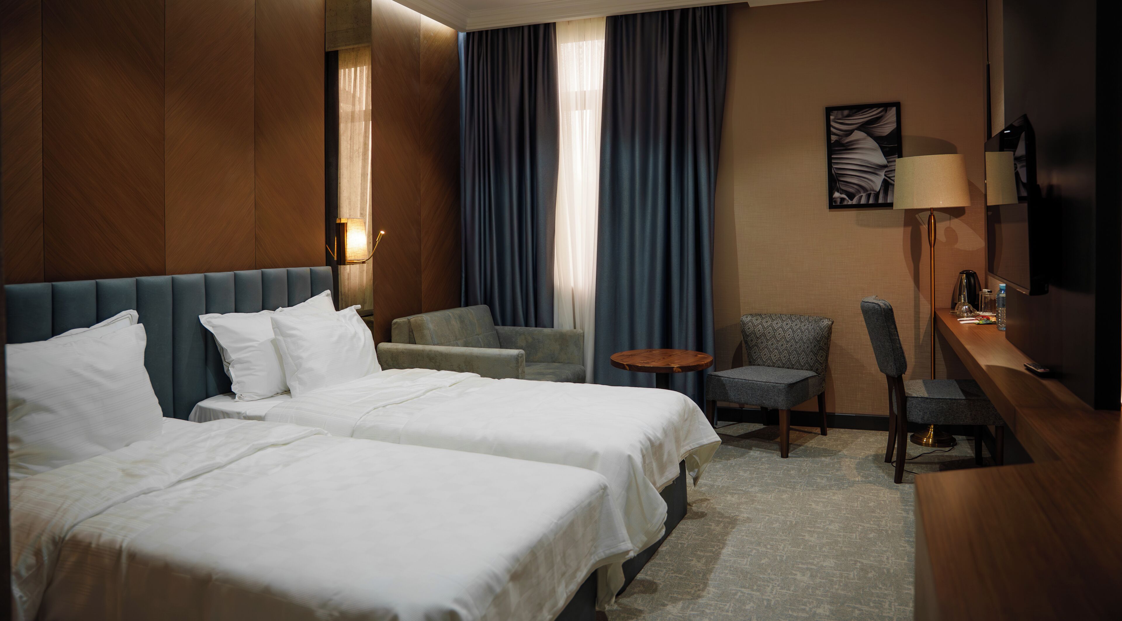 A hotel room with two beds, a desk, a chair, a small table, and a lamp.