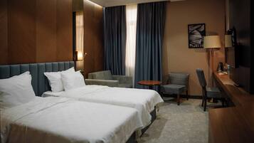 A hotel room with two beds, a desk, a chair, a small table, and a lamp.