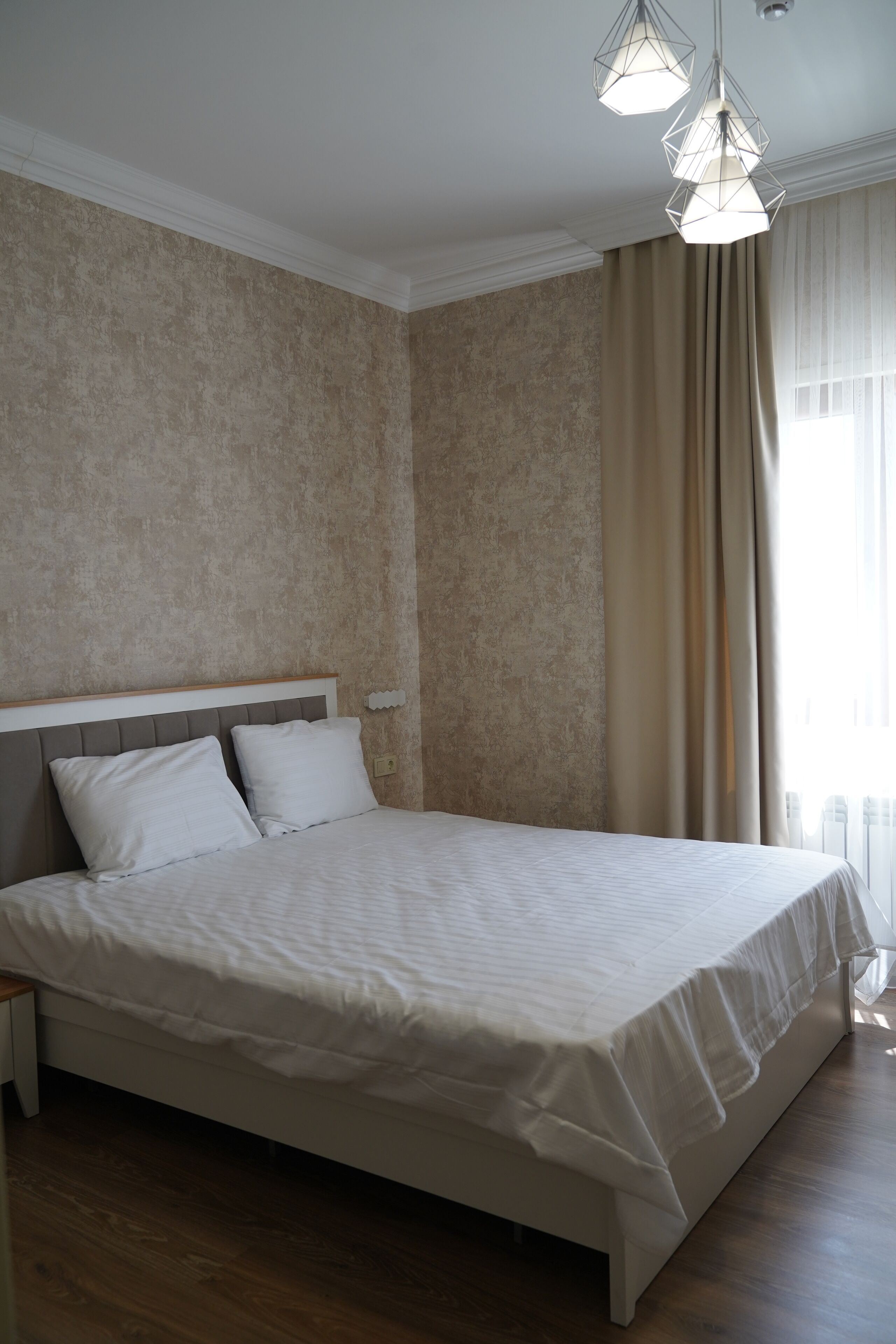 Una cama bien hecha con cabecera blanca y almohadas, una mesita de noche de madera y una ventana con cortinas.