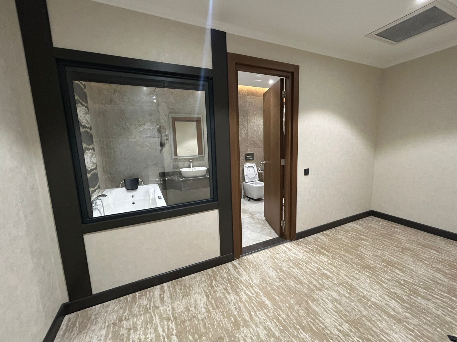 A hotel room with a large window, a bathroom visible through the door, and a carpeted floor.