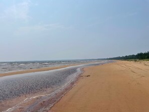 A wide sandy beach with gentle waves, a clear sky, and a distant tree line.
