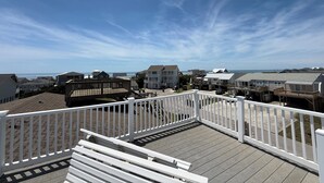 Una terraza de madera con barandas blancas, con vista a una zona residencial con casas y una playa al fondo.