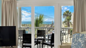 Ein Zimmer mit Fernseher, Korbstühlen, einem Balkon mit Blick auf den Strand und Palmen.