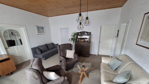 Een woonkamer met een grijze bank, een beige bank, een bruine fauteuil en een salontafel. In de hoek staat een houten kast en een plant.