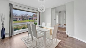 A modern dining room with a white table and chairs, a chandelier, and a view of a suburban neighborhood.