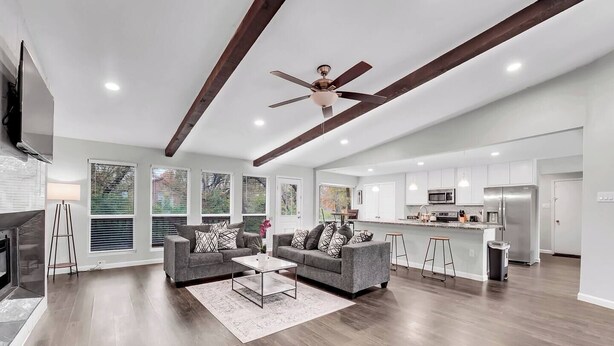A spacious living room with a fireplace, two grey sofas with patterned cushions, a coffee table, and a ceiling fan.