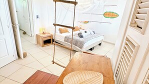Une pièce avec un lit, une table en bois, un fauteuil suspendu et une porte blanche.
