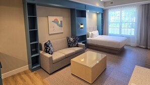 A modern hotel room with a sofa, a bed, a coffee table, and a large window with curtains.